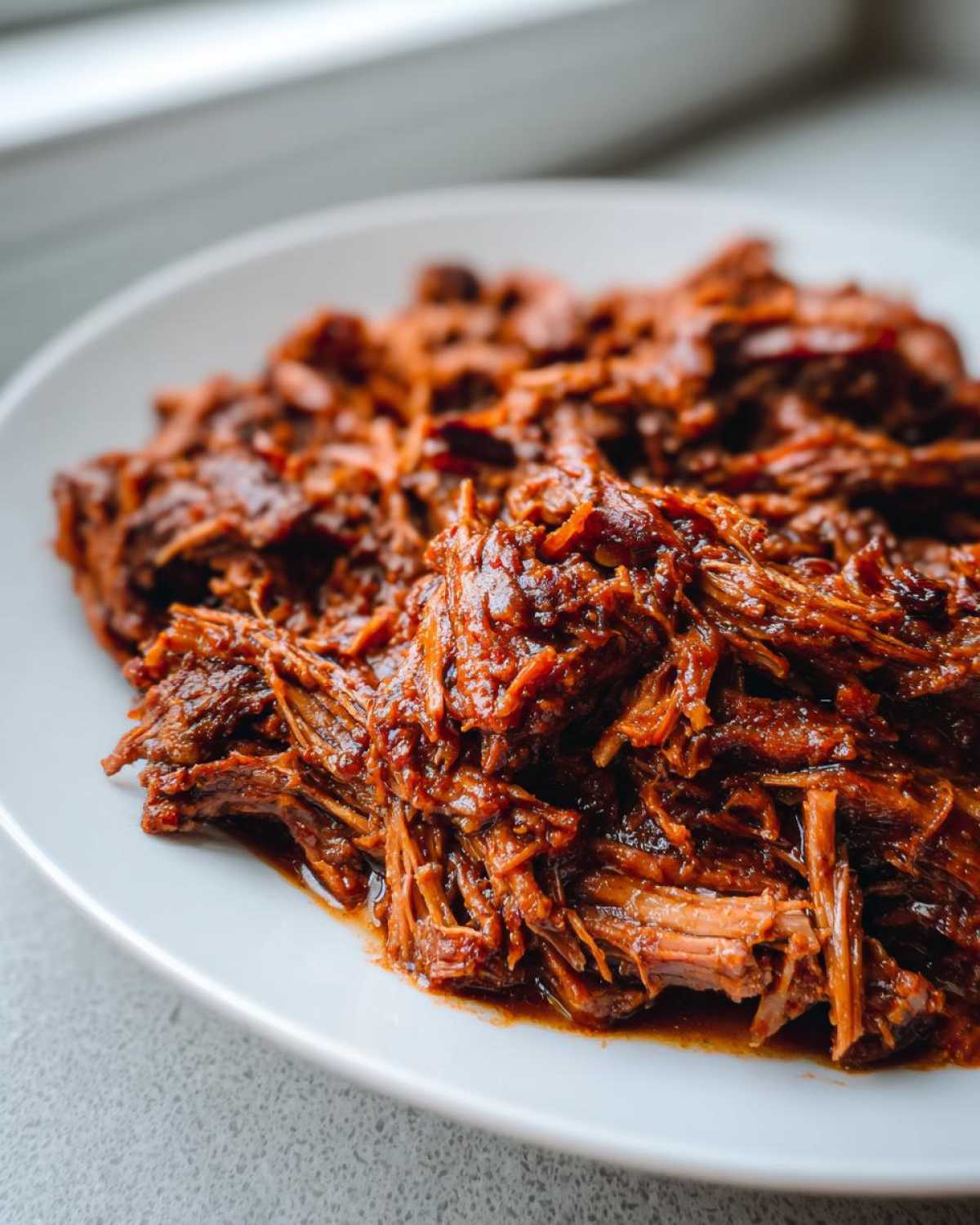 Close-up of juicy, shredded Instant Pot Beef Barbacoa coated in rich sauce, served on a white plate.