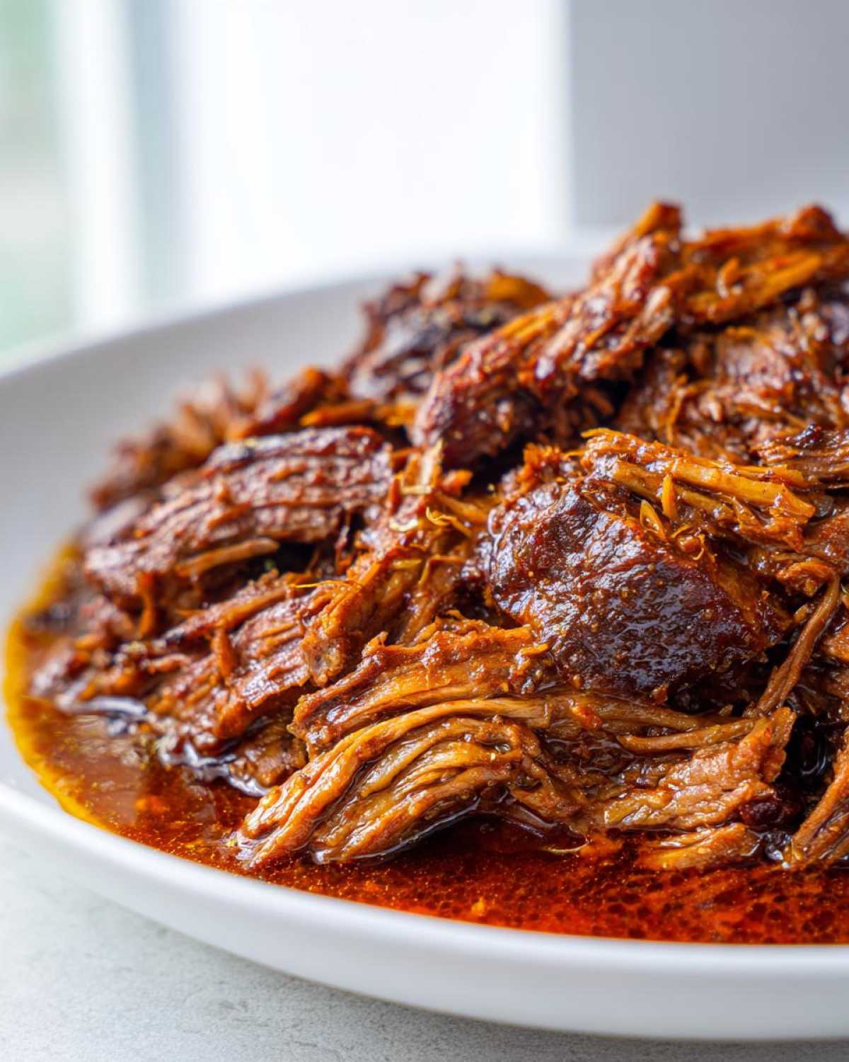 Close-up of tender, shredded Instant Pot Beef Barbacoa soaking in rich, red sauce in a white bowl.