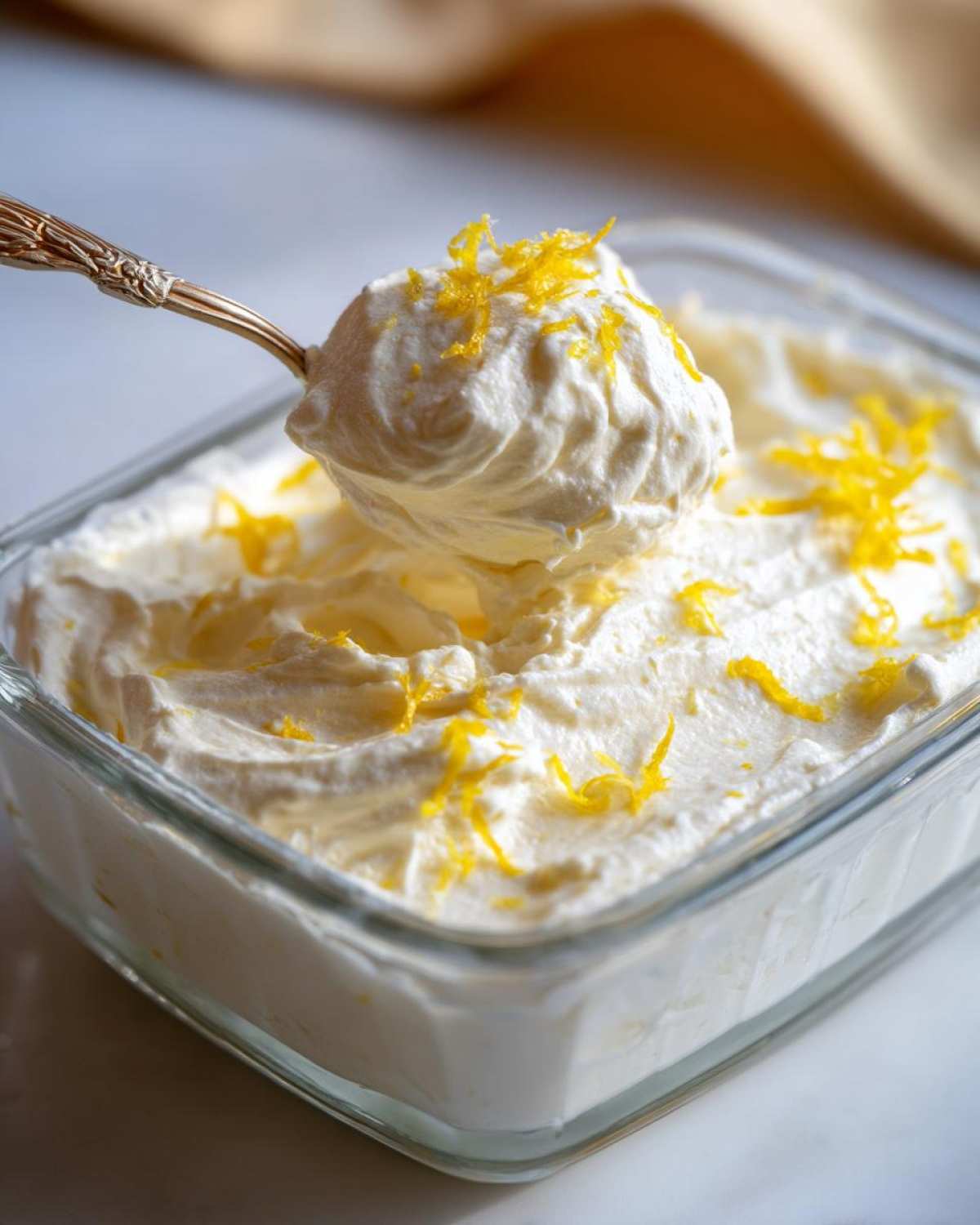 A spoonful of fluffy Keto Lemon Cheesecake Mousse topped with bright yellow lemon zest being lifted from a glass dish.