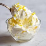 A spoonful of fluffy Keto Lemon Cheesecake Mousse topped with bright yellow lemon zest being lifted from a clear glass bowl.