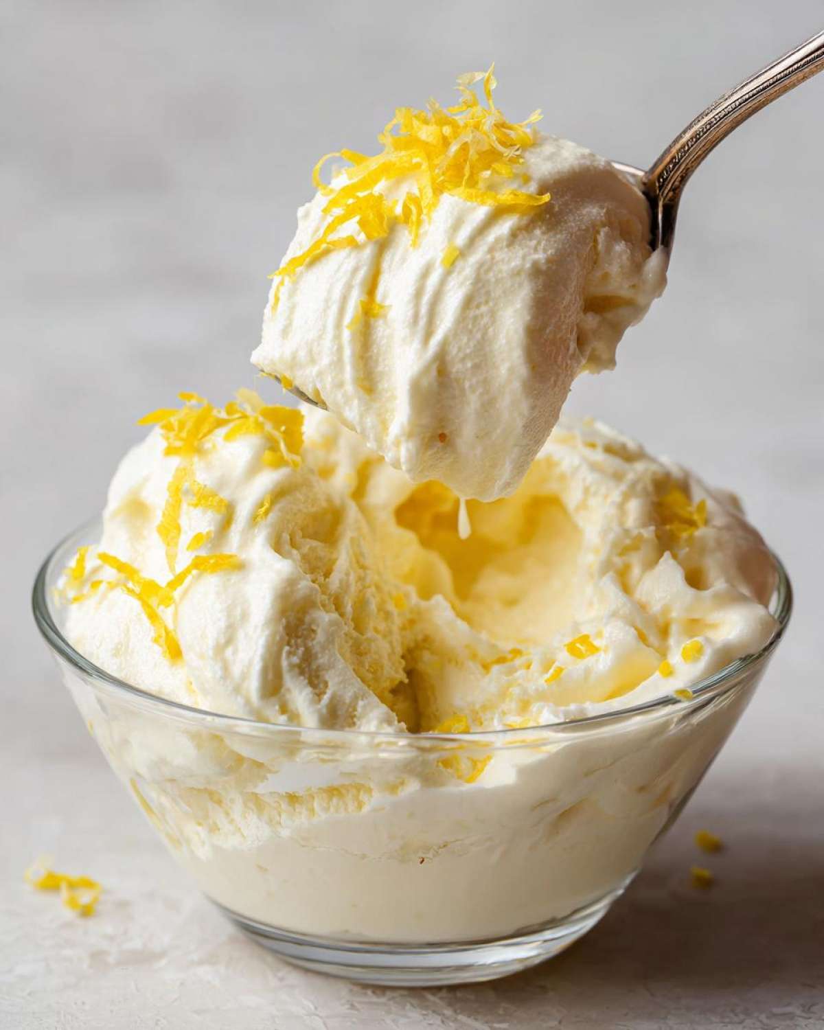 A spoonful of fluffy Keto Lemon Cheesecake Mousse being lifted from a glass bowl, topped with bright yellow lemon zest.