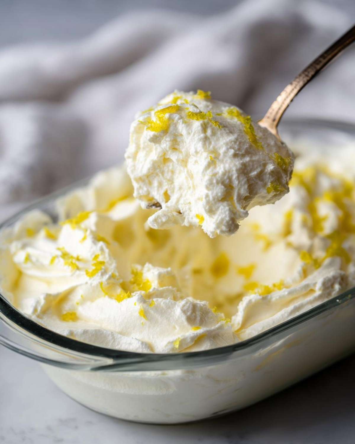 A spoonful of fluffy Keto Lemon Cheesecake Mousse topped with bright yellow lemon zest being lifted from a glass dish.