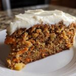 A close-up of a thick slice of Moist Carrot Cake with Cream Cheese Frosting showing visible carrot shreds and nuts.