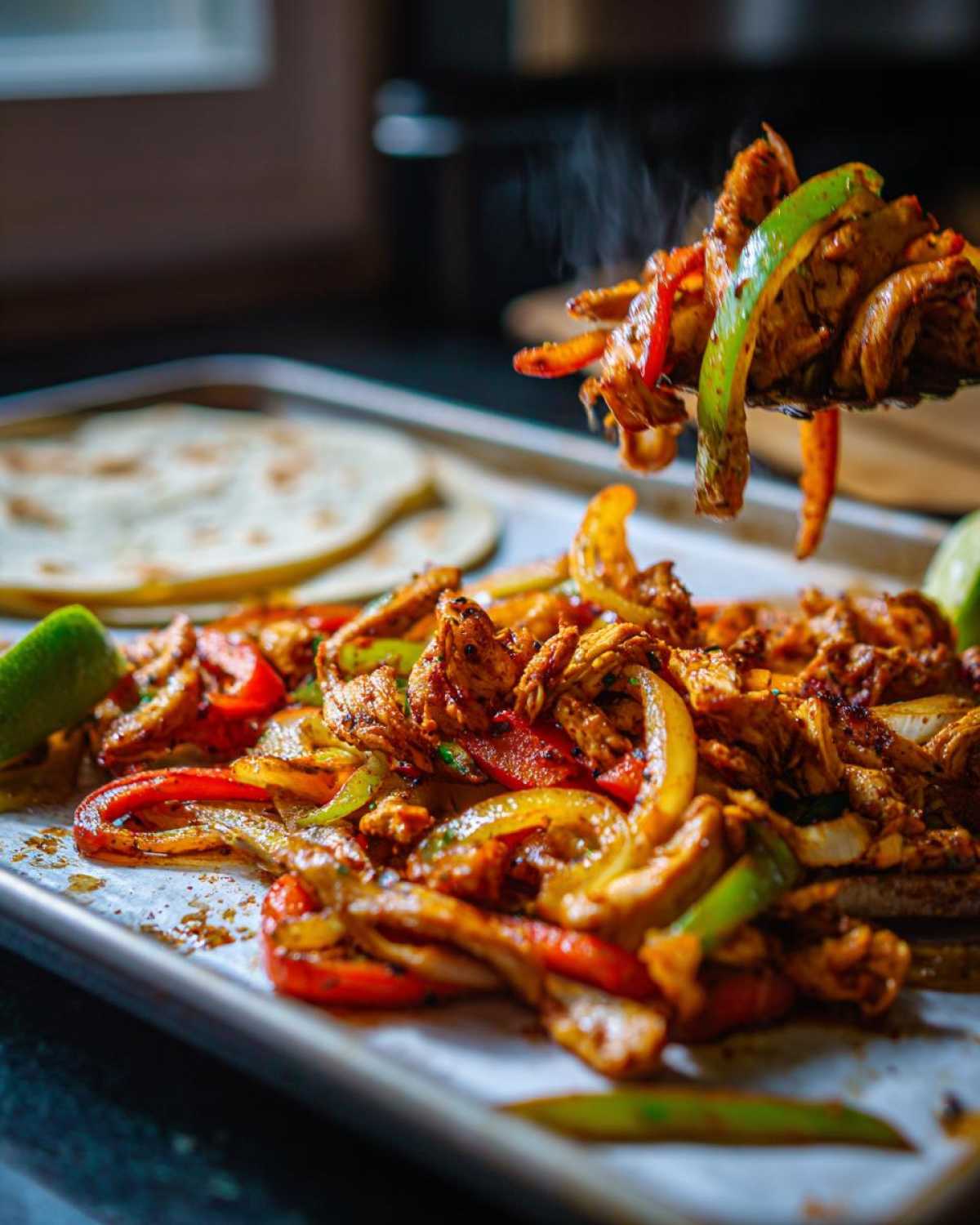 A spatula lifts hot, steaming One-Pan Chicken Fajitas with peppers and onions off a baking sheet next to tortillas.