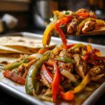 A wooden spoon lifts sizzling One-Pan Chicken Fajitas with Peppers and Onions from a platter next to warm tortillas.