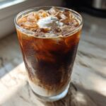 A tall glass of cold Iced Mocha showing layers of milk, coffee, ice cubes, and a dollop of whipped cream.