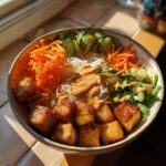 A vibrant Spring Roll Bowl with Peanut Sauce featuring crispy tofu, rice noodles, shredded carrots, and sliced cucumbers.