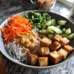 A vibrant Spring Roll Bowl with Peanut Sauce featuring vermicelli noodles, crispy tofu cubes, shredded carrots, cucumbers, and fresh herbs.