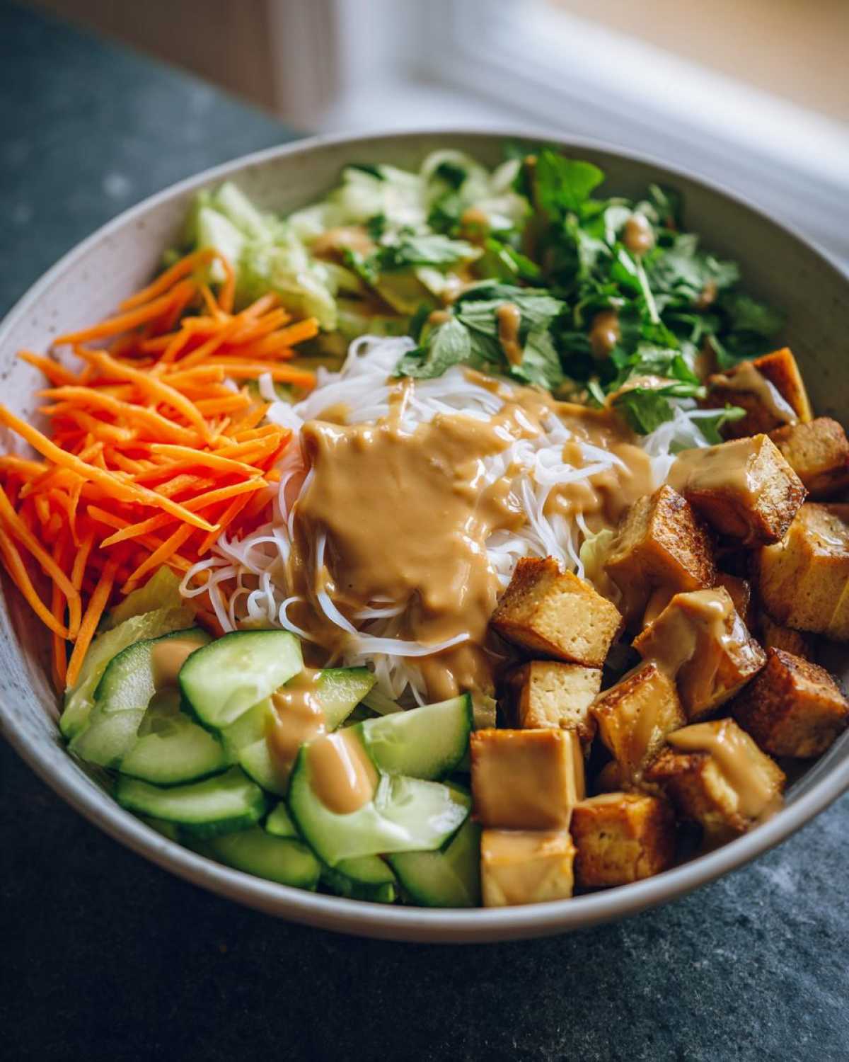 Close-up of a vibrant Spring Roll Bowl with Peanut Sauce, featuring rice noodles, crispy tofu, shredded carrots, and cucumbers.
