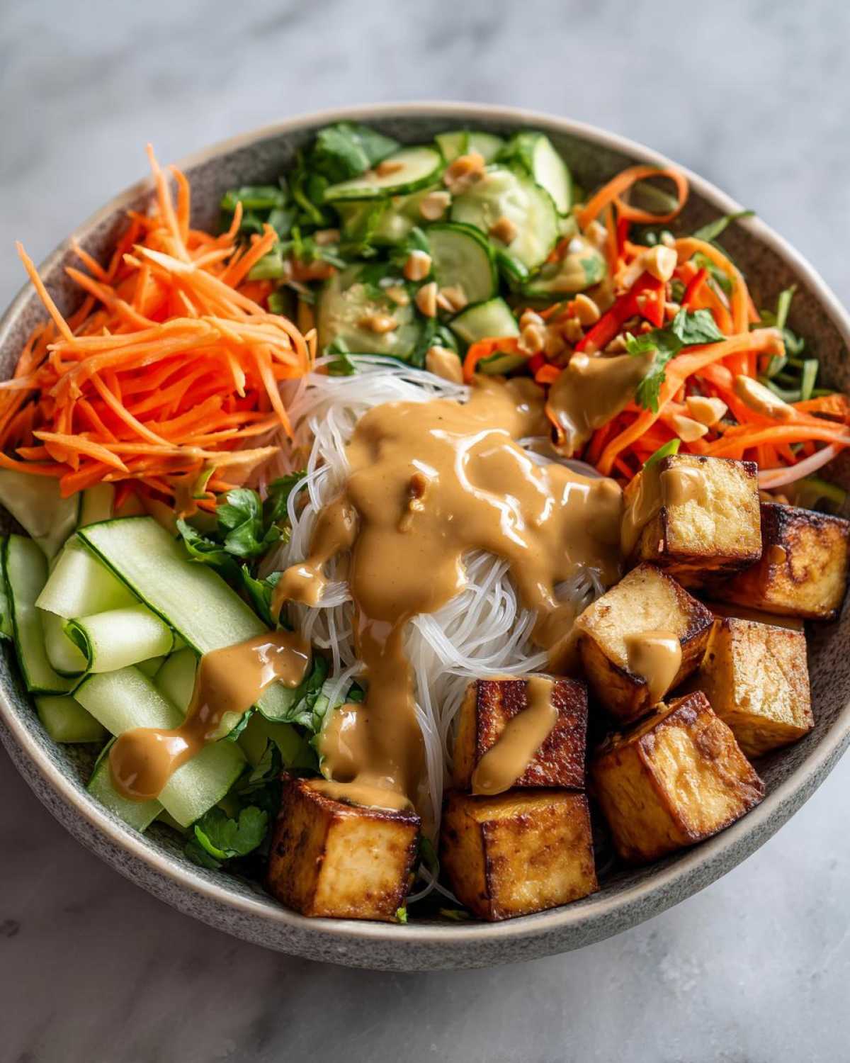 A vibrant Spring Roll Bowl with Peanut Sauce featuring rice noodles, crispy tofu cubes, shredded carrots, and sliced cucumbers.