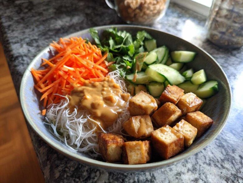 A vibrant Spring Roll Bowl with Peanut Sauce featuring vermicelli noodles, crispy tofu cubes, shredded carrots, cucumbers, and fresh herbs.