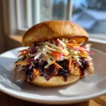 A close-up of a Sticky BBQ Chicken Sandwich with pulled chicken dripping with sauce and topped high with colorful crunchy slaw.