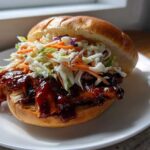 A close-up of a Sticky BBQ Chicken Sandwich with crunchy coleslaw piled high on a soft bun, served on a white plate.