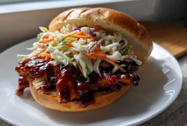 A close-up of a Sticky BBQ Chicken Sandwich with crunchy coleslaw piled high on a soft bun, served on a white plate.