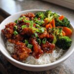 A close-up of a Sticky Orange Chicken Bowl with rice, steamed broccoli, carrots, and green onions.