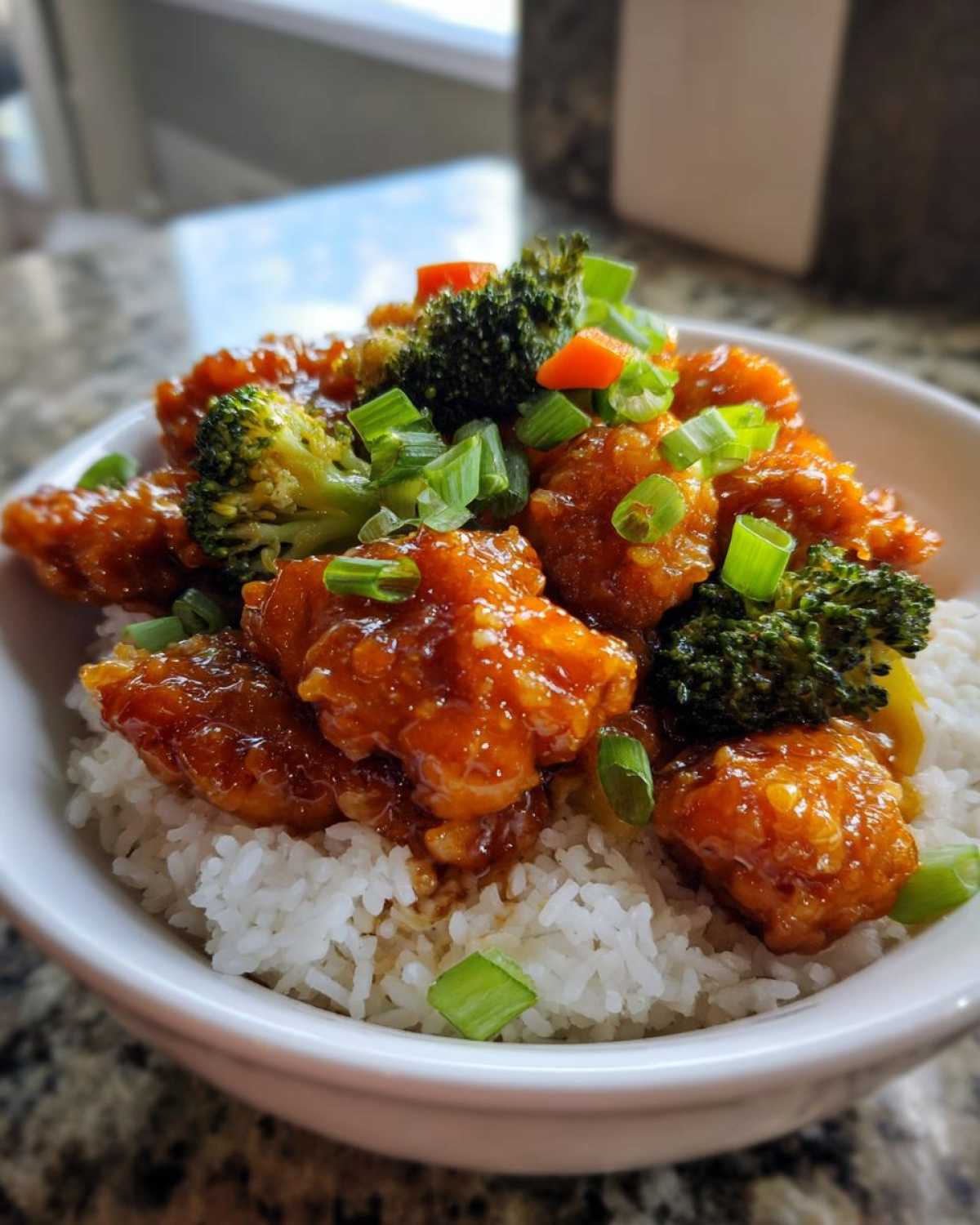 Close-up of a Sticky Orange Chicken Bowl featuring glazed chicken pieces over white rice, topped with broccoli, carrots, and green onions.