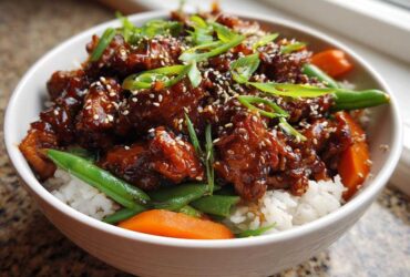 A close-up of a white bowl filled with Sticky Teriyaki Chicken Bowl, featuring glazed chicken over white rice, green beans, carrots, and sesame seeds.
