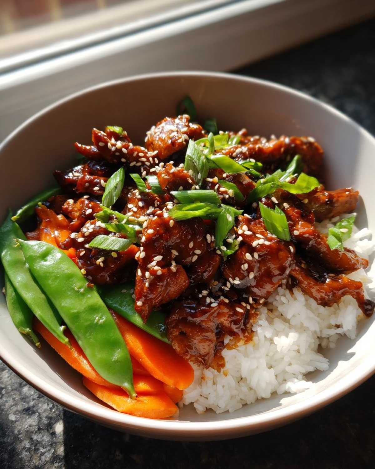 A close-up of a Sticky Teriyaki Chicken Bowl featuring glazed chicken pieces over white rice, served with bright green snow peas and sliced carrots.