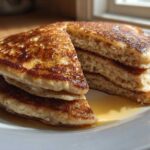 A stack of fluffy Vegan Banana Oat Pancakes, cut to show the interior texture, drizzled with maple syrup on a white plate.