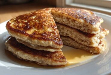A stack of fluffy Vegan Banana Oat Pancakes, cut to show the interior texture, drizzled with maple syrup on a white plate.