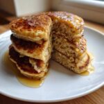 A stack of fluffy Vegan Banana Oat Pancakes cut in half, drizzled generously with maple syrup on a white plate.